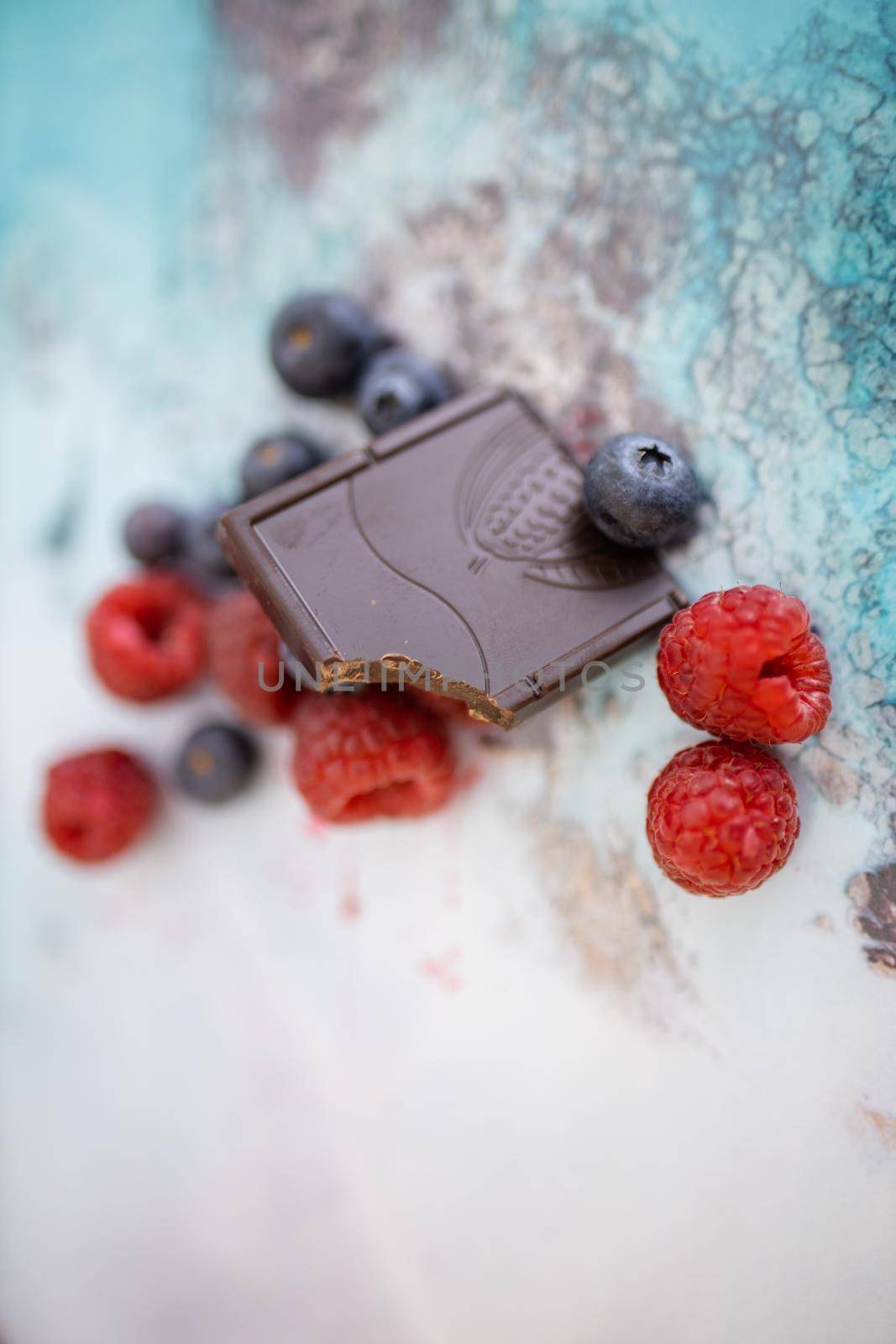 Bitten chocolate bar leaning against raspberries and blueberries on colorful placemat. Dark cacao bar and fruit on table from above. Fresh berries and chocolate