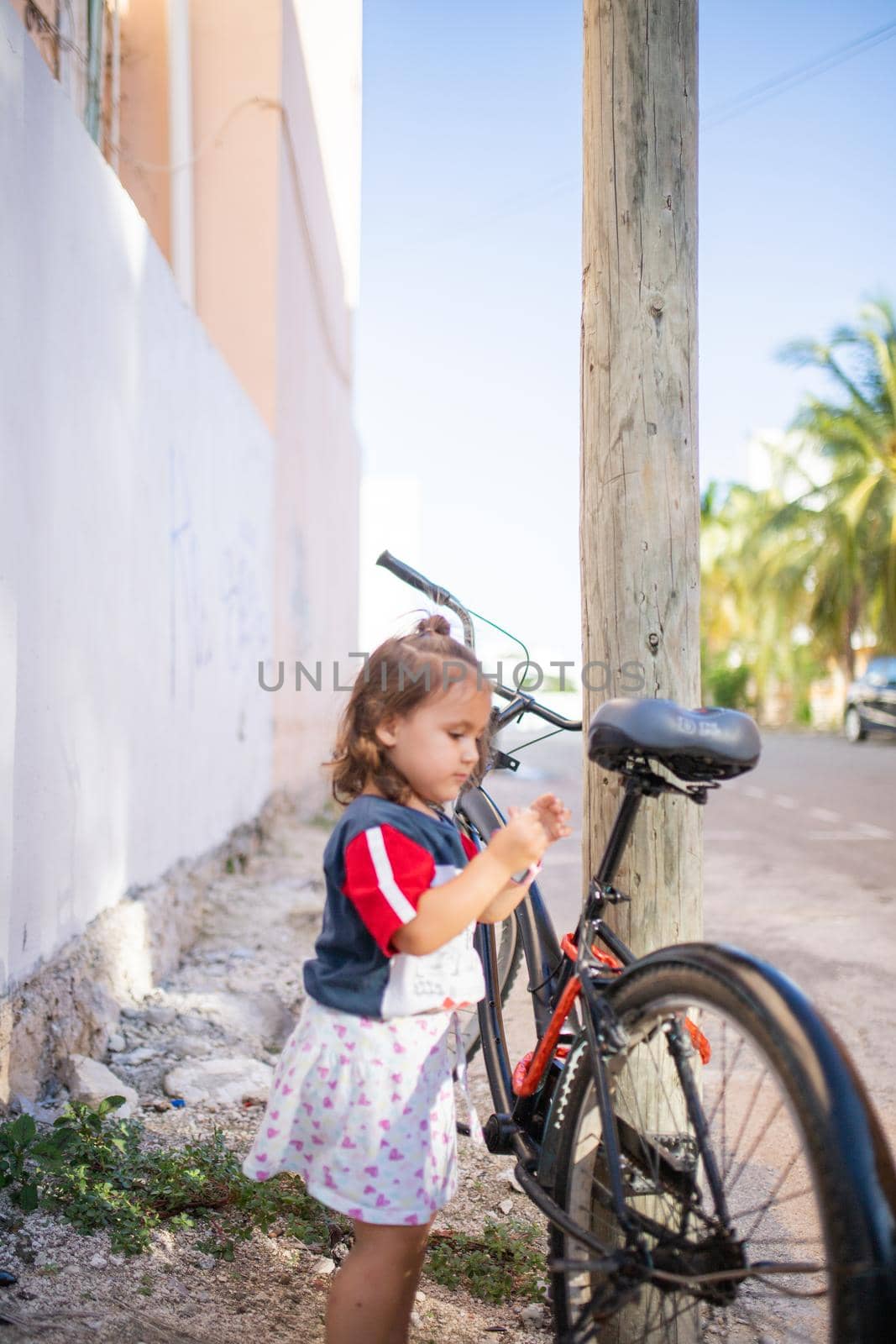 Adorable little girl playing next to bicycle that leans against wooden lamppost. Portrait of cute young child standing next to black bike in the street. Tropical summer vacations
