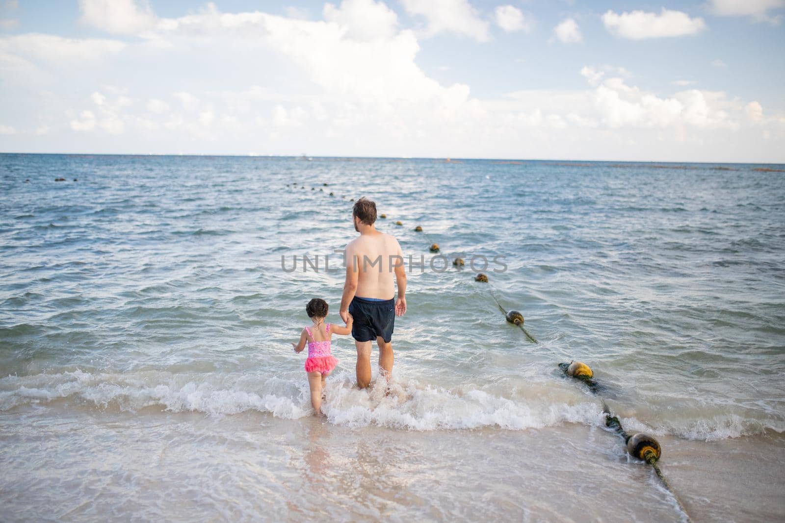 Father and young daughter holding hands as they walk on the beach and into the water. Man and little girl standing on the beach with beautiful sea in the distance. Tropical summer vacations