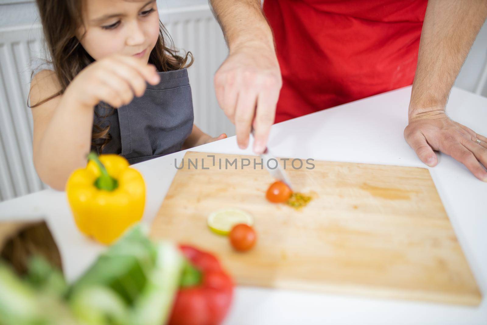 Happy little girl and her father slicing cherry tomatoes on cutting board. Male hands helping young child cutting vegetables on wooden board. Daughter-father cooking together