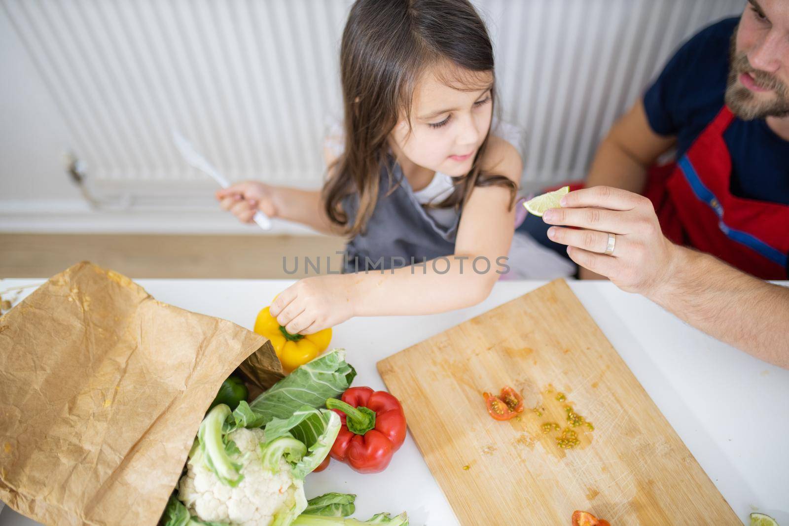 Father at white table giving a lemon slice to happy little daughter. Man and young child cutting vegetables on wooden board. Daughter-father cooking together