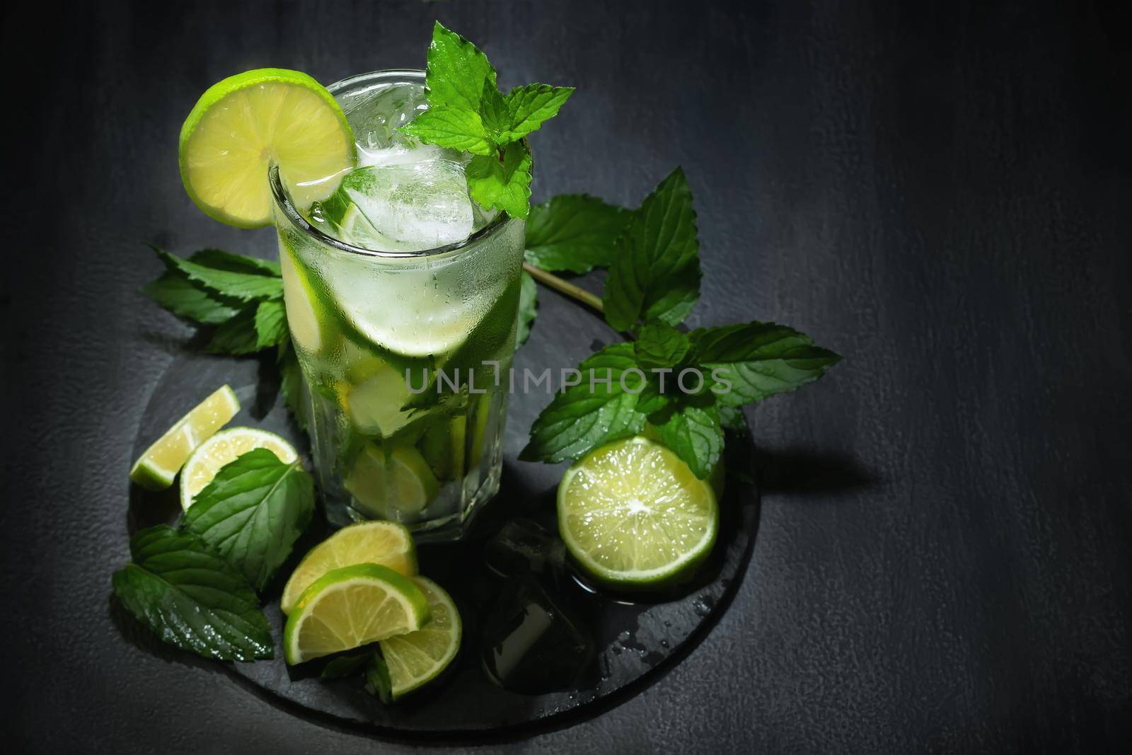 Fresh homemade mojito cocktail in a tall glass with lime, mint and ice on a black table, copy space by galsand