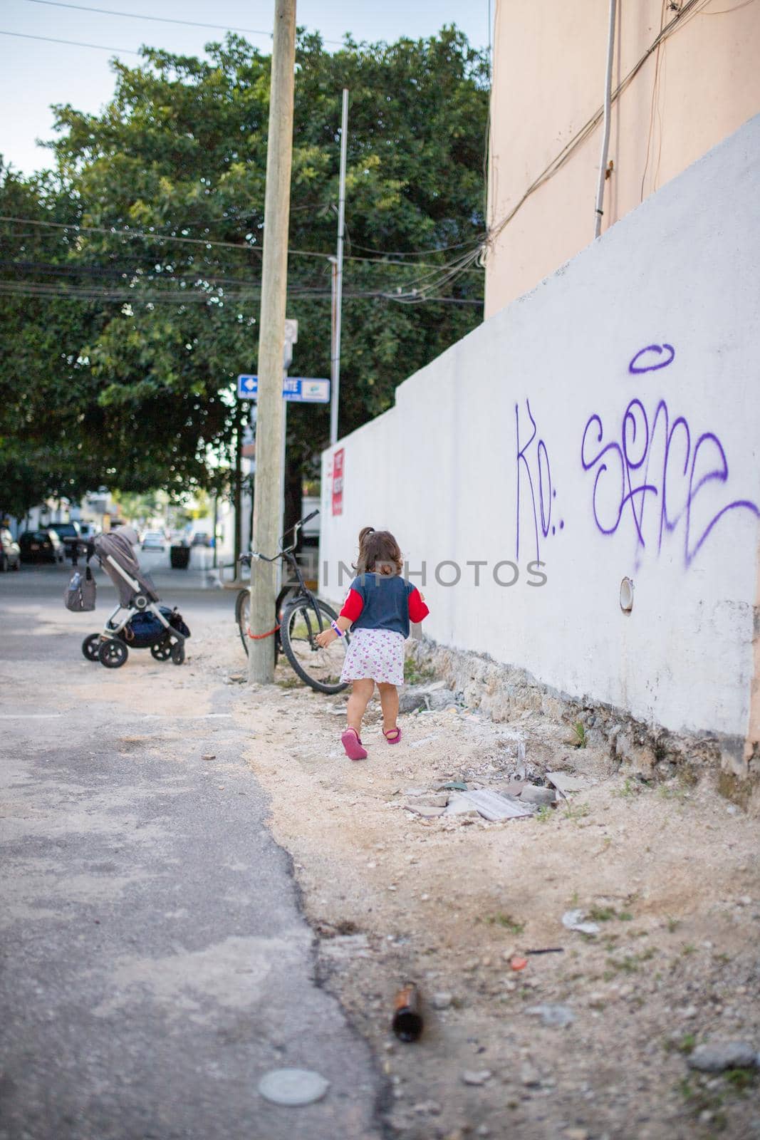 Adorable little girl running towards bicycle that leans against wooden lamppost. Portrait of cute young child wearing white skirt in the street. Tropical summer vacations