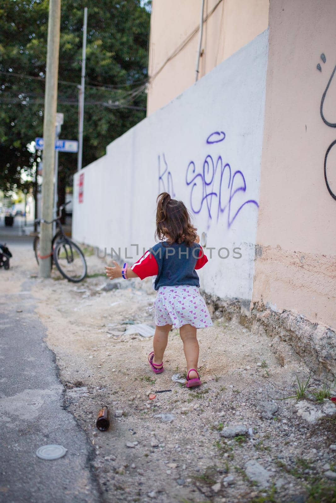 Adorable little girl running towards bicycle that leans against wooden lamppost. Portrait of cute young child wearing white skirt in the street. Tropical summer vacations