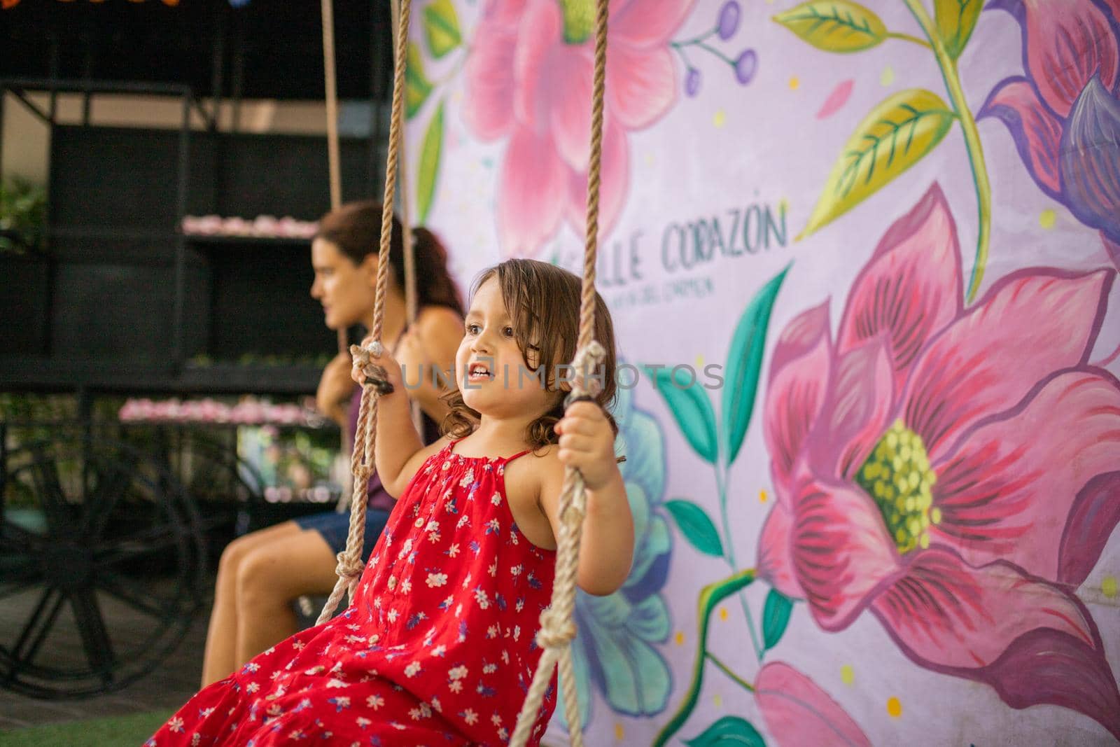 Adorable little girl and woman sitting in rope swings in front of wall with painted flowers and the word heart. Cute young child wearing red dress and having fun on swing. Daughter-mother fun day
