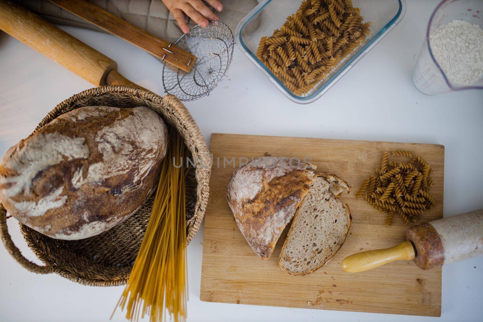 Bread, pasta, flour, and wooden cookware on white table from above. Ingredients and carbohydrates on cutting board and a basket. Healthy meal preparation