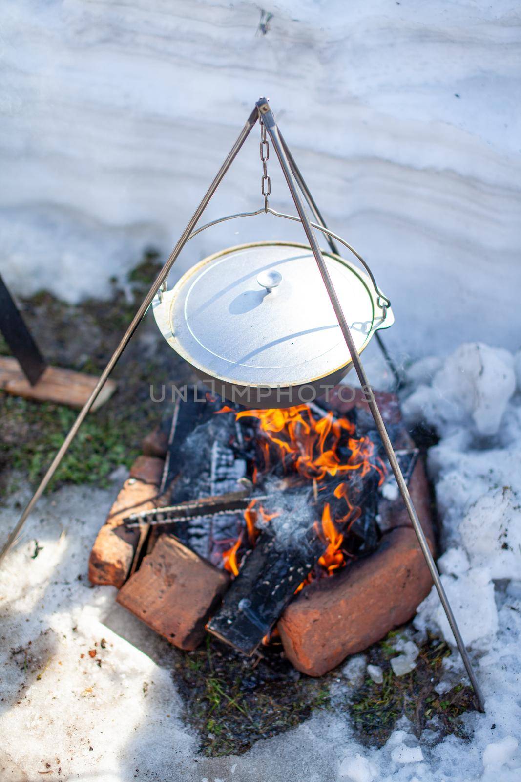 Over the fire hangs a pot in which to cook food. On a hook on a tripod, steam comes out of the pan. Winter Camping outdoor cooking