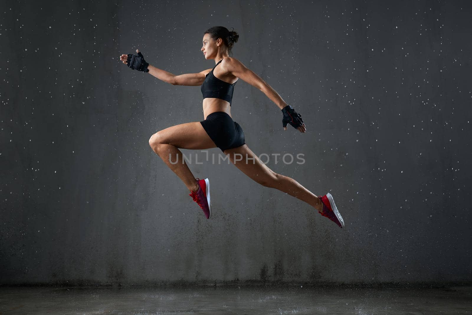 Isolated portrait of athletic woman jumping in hall under rain, loft gray interior. Side view of wet female bodybuilder in sports underwear doing running pose in air during jump. Concept of sport.