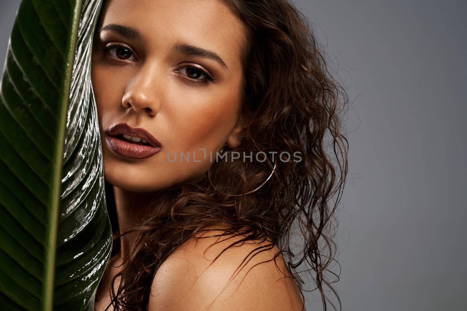 Close up of beautiful naked female model looking at camera. Crop portrait of young brunette woman with perfect makeup posing with mouth open, holding big green leaf, isolated on gray.