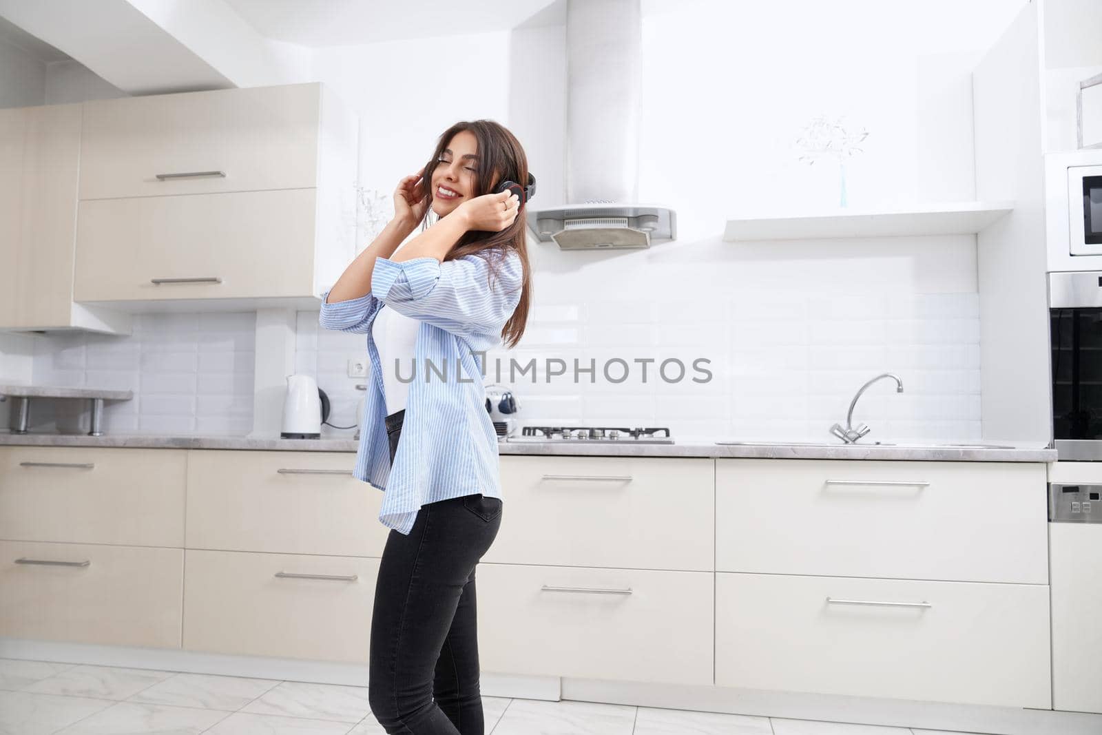 Smiling woman standing and listening music in headphone at home. Concept of enjoying free time in kitchen.