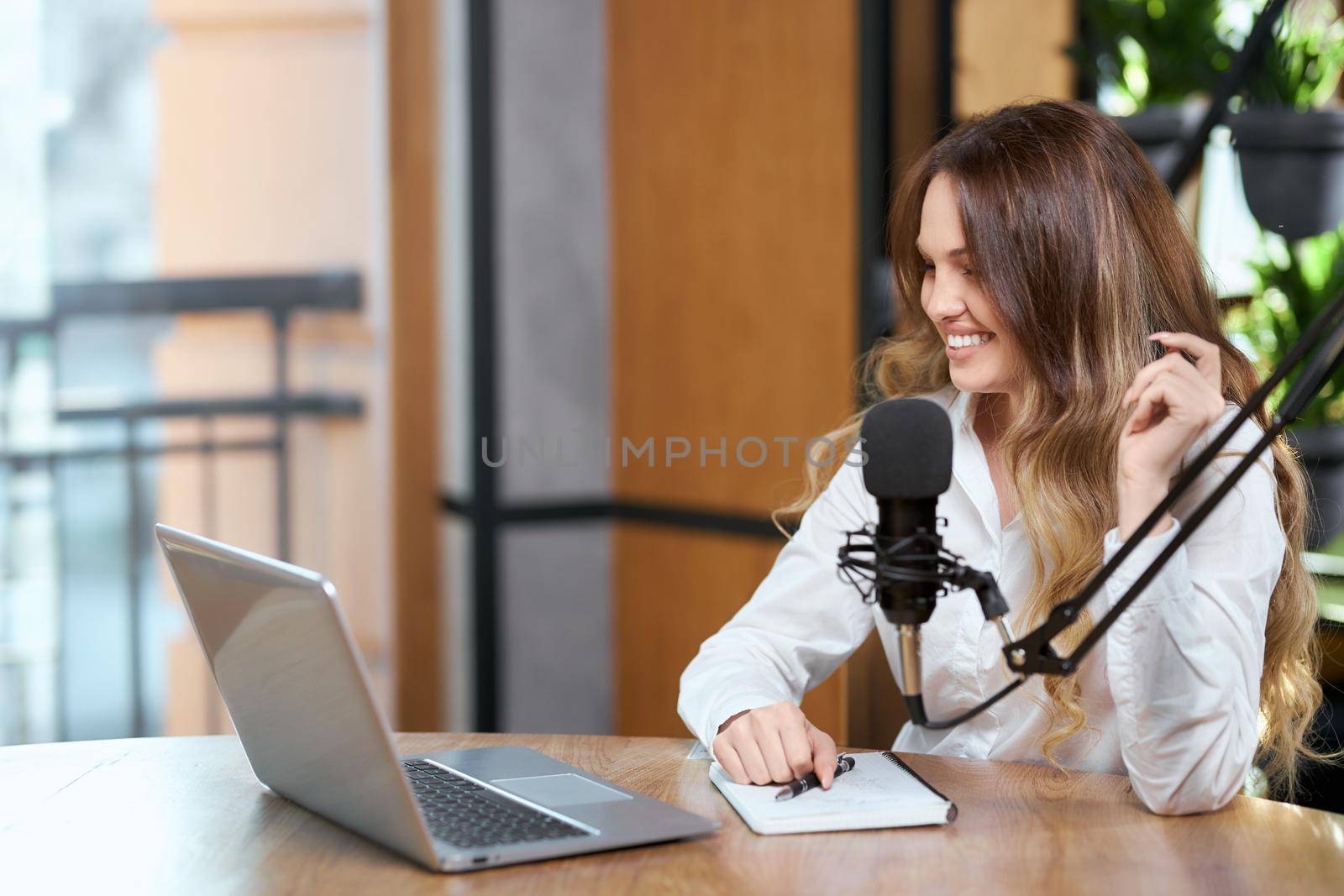 Side view of smiling beautiful woman in white shirt communicating with microphone online by laptop in cafe. Concept of process talking with followers online with good mood. 