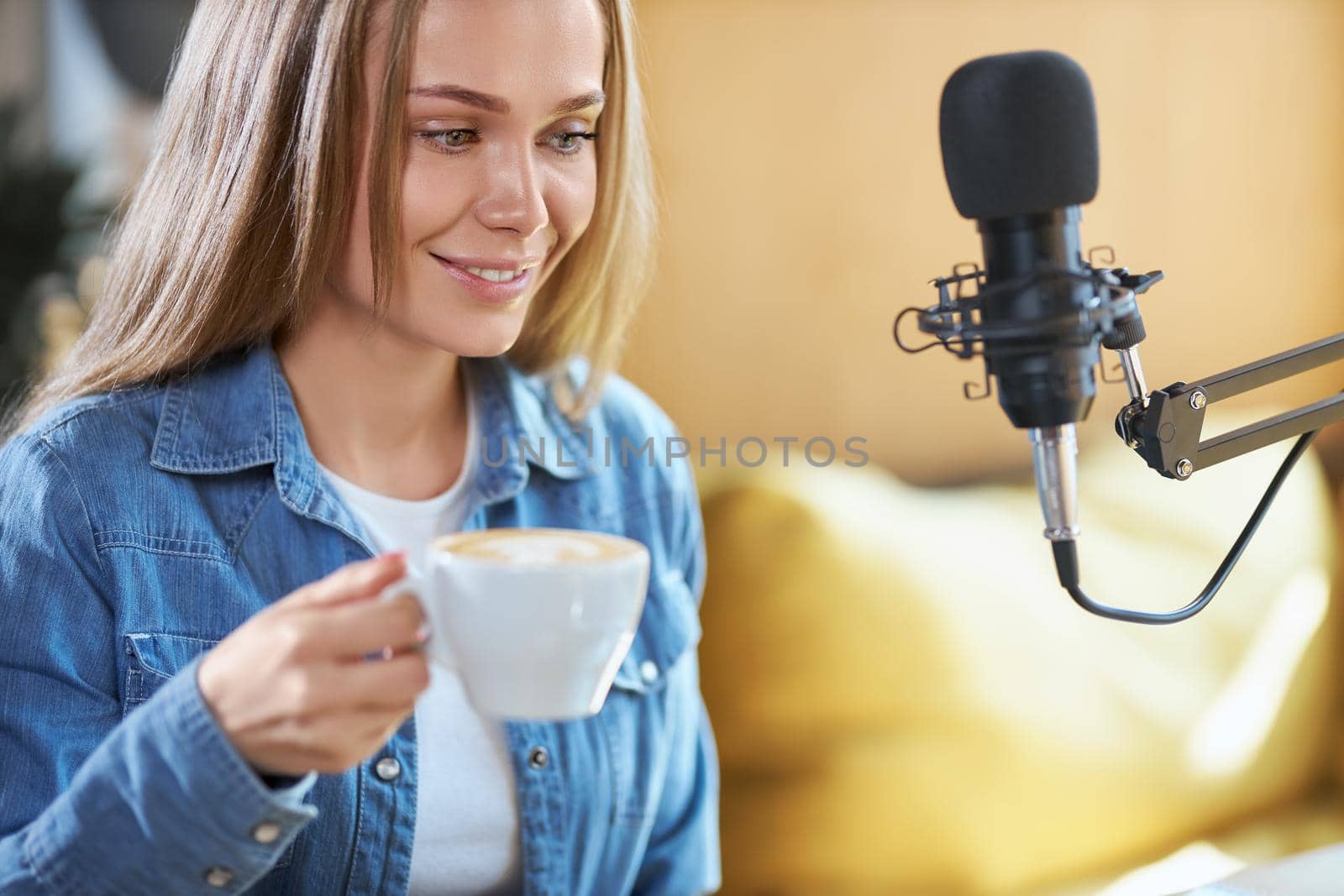 Side view of smiling beautiful young woman holding cup coffee and preparing for broadcast on the radio. Concept of process communicating or telling about interesting topics. 