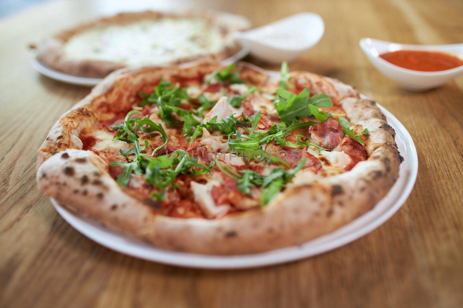 Close up of tasty pizza with ham, rucola and another pizza with different cheeses and with red sauce. Concept of delicious lunch or dinner with tasty pizza on white plate in cafe.