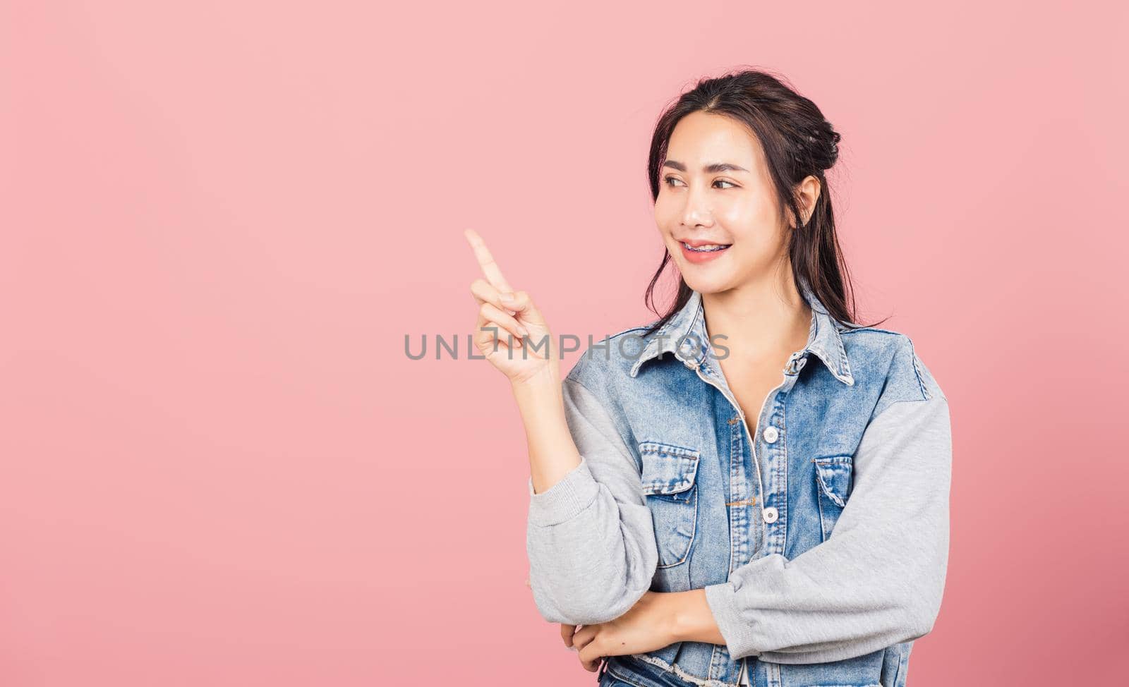 Happy Asian portrait beautiful cute young woman standing smiling indicate finger empty space looking to space, studio shot isolated on pink background, Thai female pointing index out with copy space