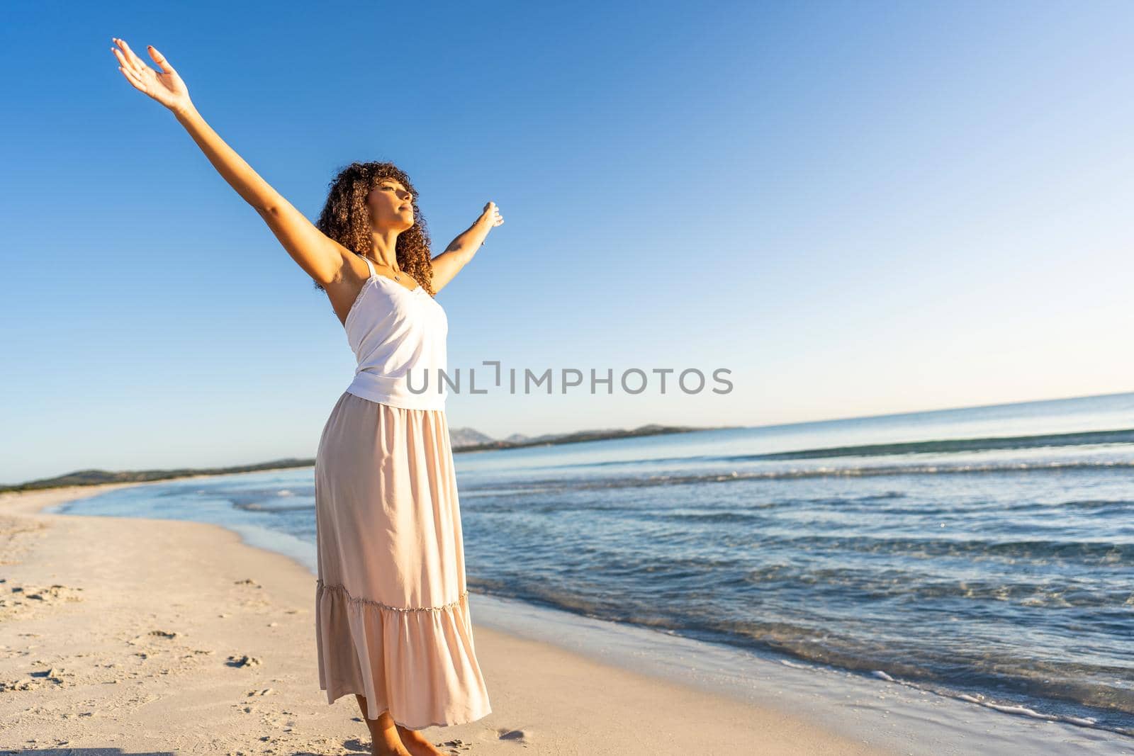 Beautiful twenty year old African American woman with arms outstretched in adoration of nature standing on sand looking at sun just rising over the ocean sea. Concept of purity, love and spirituality