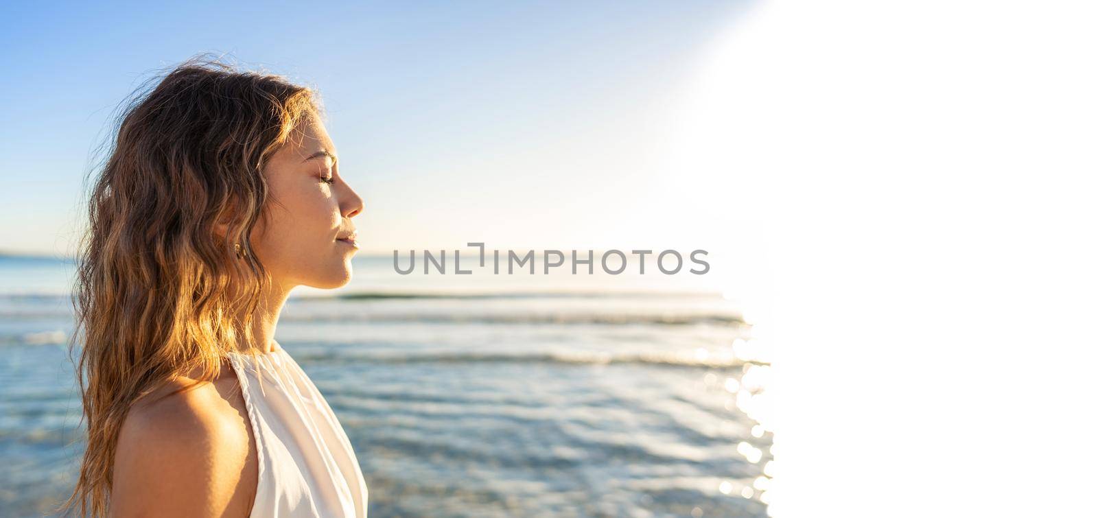 Profile portrait of young beautiful Caucasian blond woman with closed eyes in a white boho dress getting kissed by sunlight at sunset or dawn at beach with ocean sea in background and white copy space