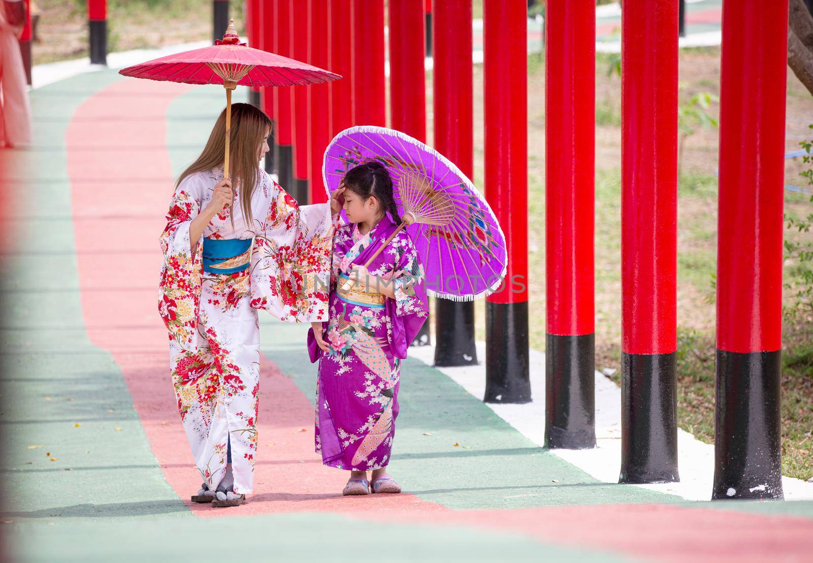  woman and little girl in kimono holding umbrella walking into at the shrine red gate, in Japanese garden.
