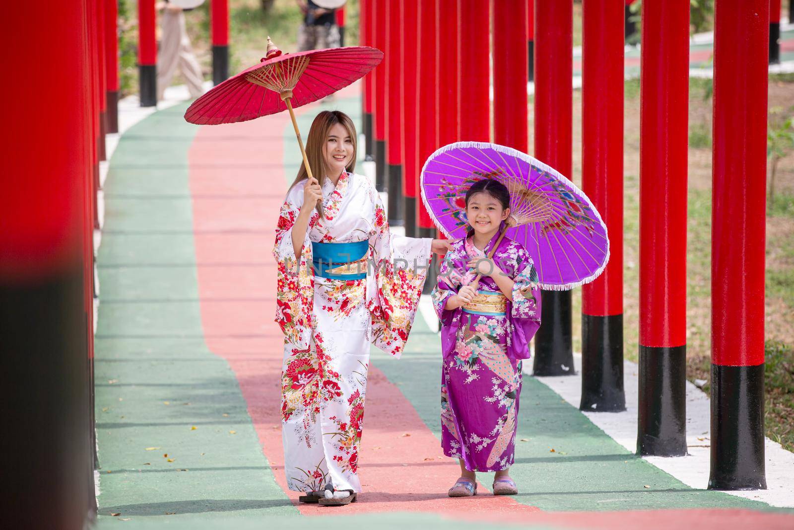  woman and little girl in kimono holding umbrella walking into at the shrine red gate, in Japanese garden.