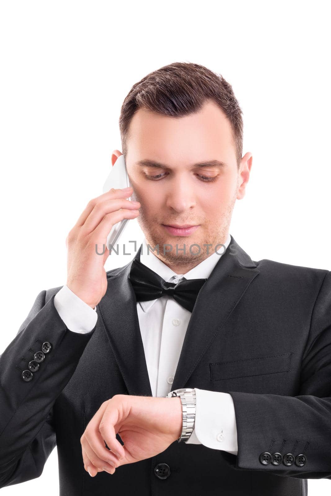 Close up portrait of a handsome serious man in black suit with bow tie talking on mobile phone while looking at his wristwatch, isolated on white background. Late concept.