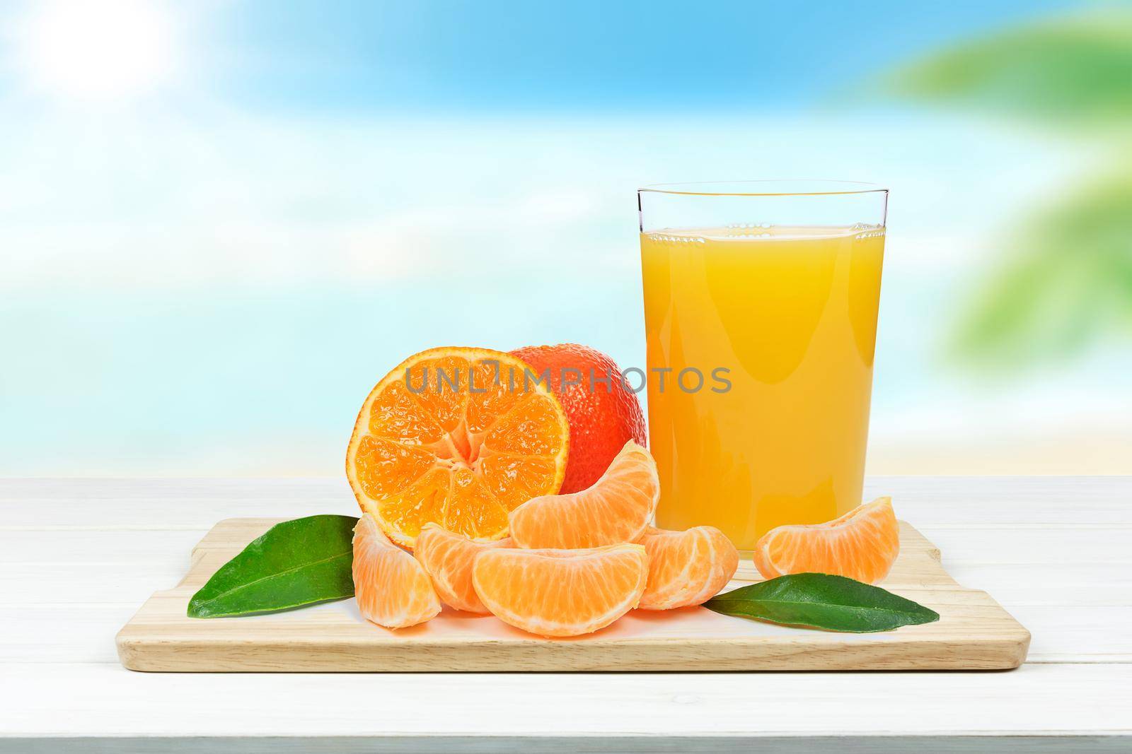 Glass of fresh orange juice and orange or tangerine fruits with leaves on blurred background of a tropical beach (mixed)