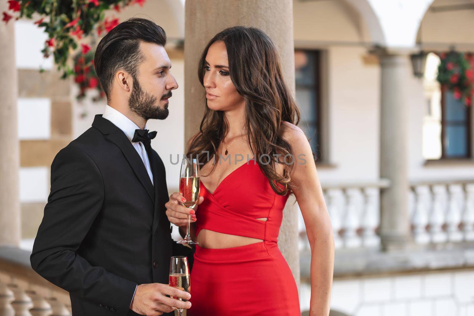 Elegant couple of lovers with a glass of wine or champagne standing on a balcony during luxury party.