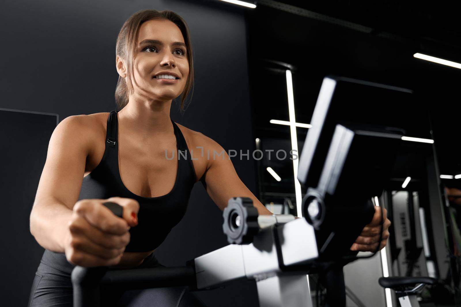 Close up of cute young brunette woman in black sportswear doing workout on exercise bike. Concept of process workout in modern gym or fitness studio.