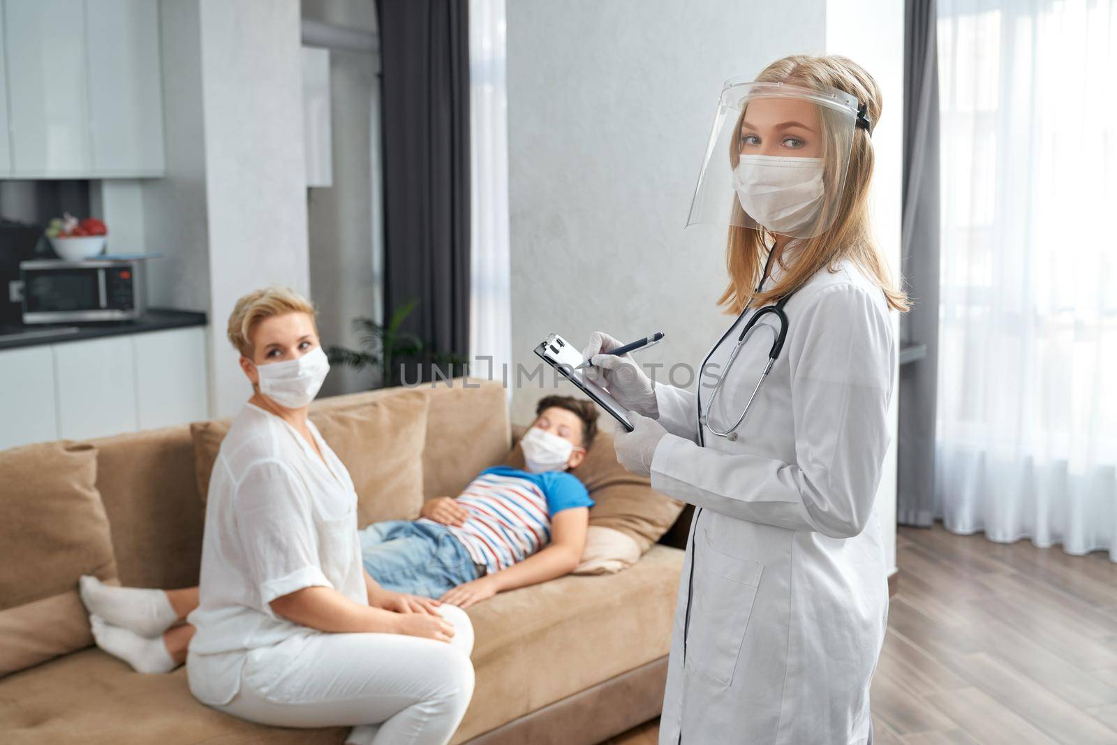 Qualified female pediatrician in medical uniform and mask writing some information on clipboard while pleasant woman sitting on couch near her sick son. Domestic checkup.