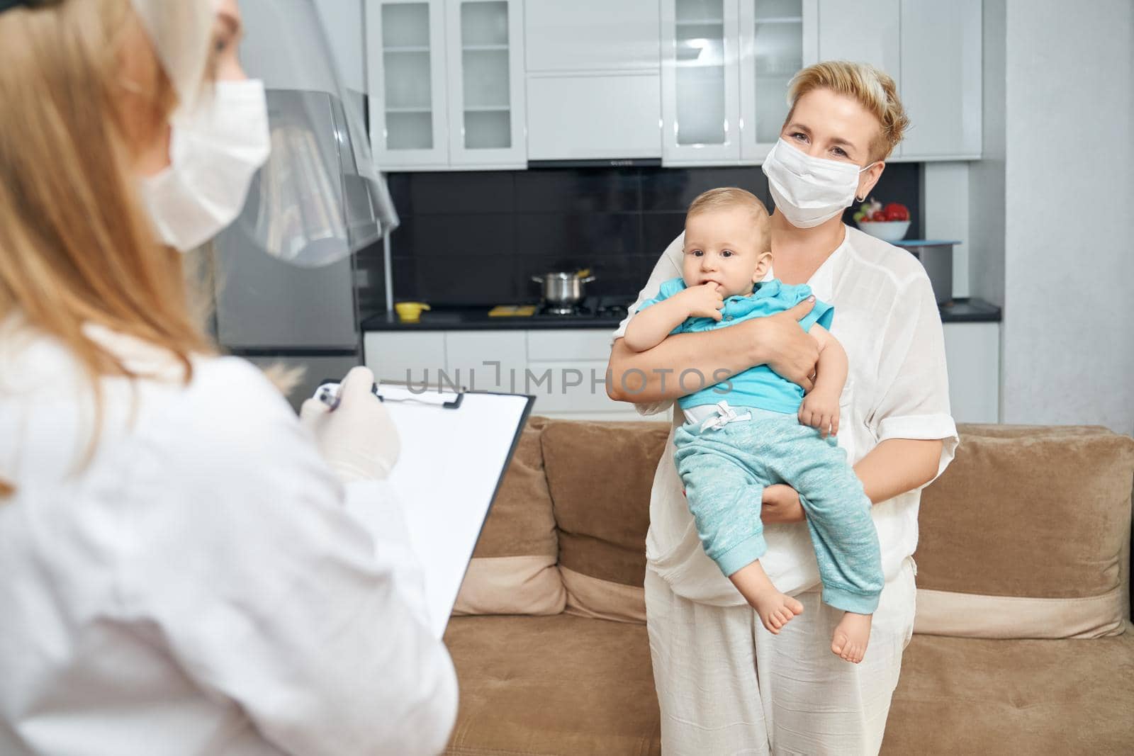 Pretty mother in face mask carrying little boy on hands while competent doctor in medical uniform writing on clipboard. Health treatment for children.