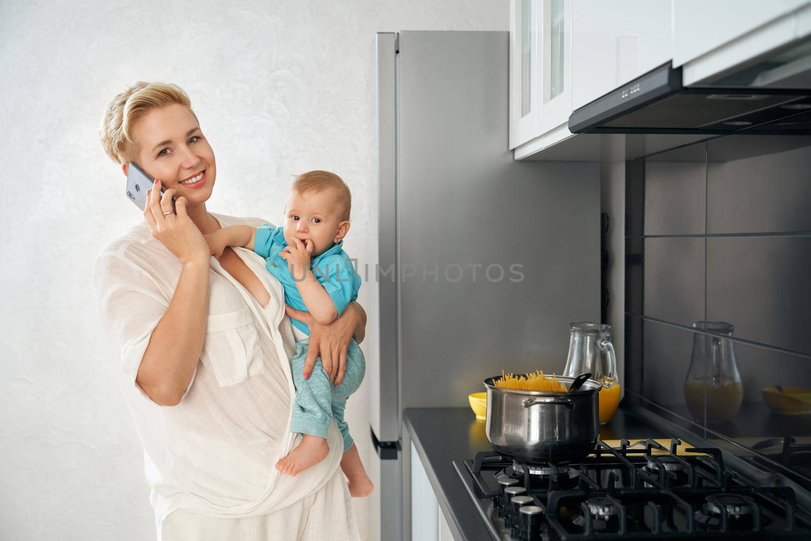 Woman talking on mobile, cooking dish and carrying baby by SerhiiBobyk