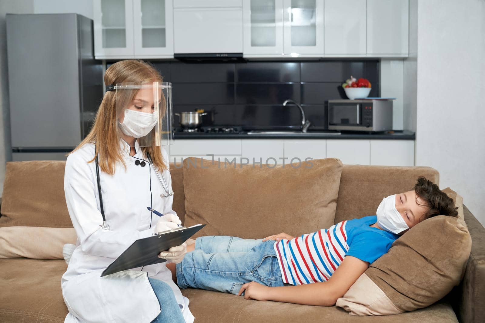 Competent pediatrician in uniform, protective mask and gloves visiting sick teenager boy at home. Female doctor writing on clipboard during health treatment of child.