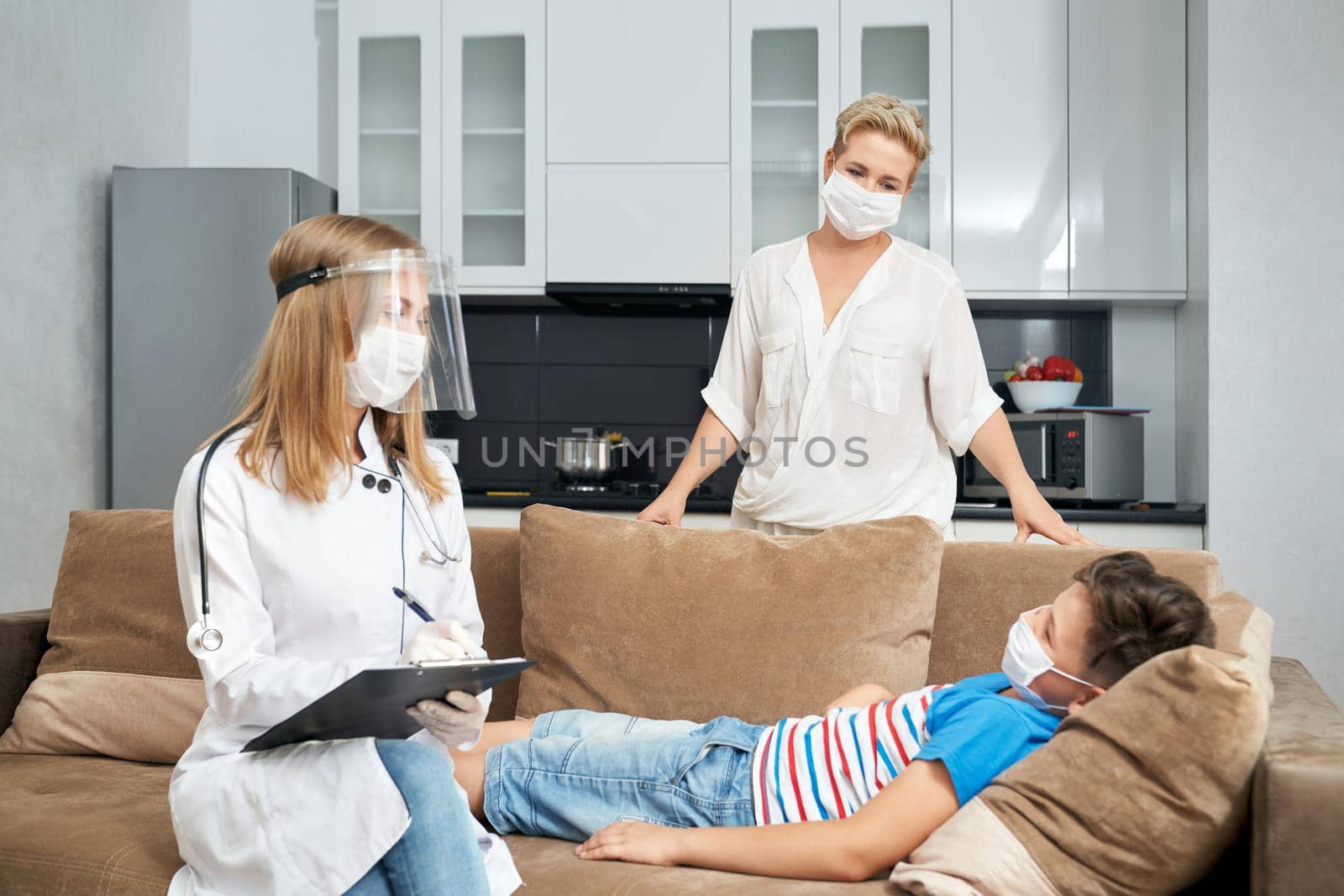 Sick teenager boy lying on cozy sofa while mother standing bear and doctor writing on clipboard. People wearing protective mask during pandemic. Children health care.