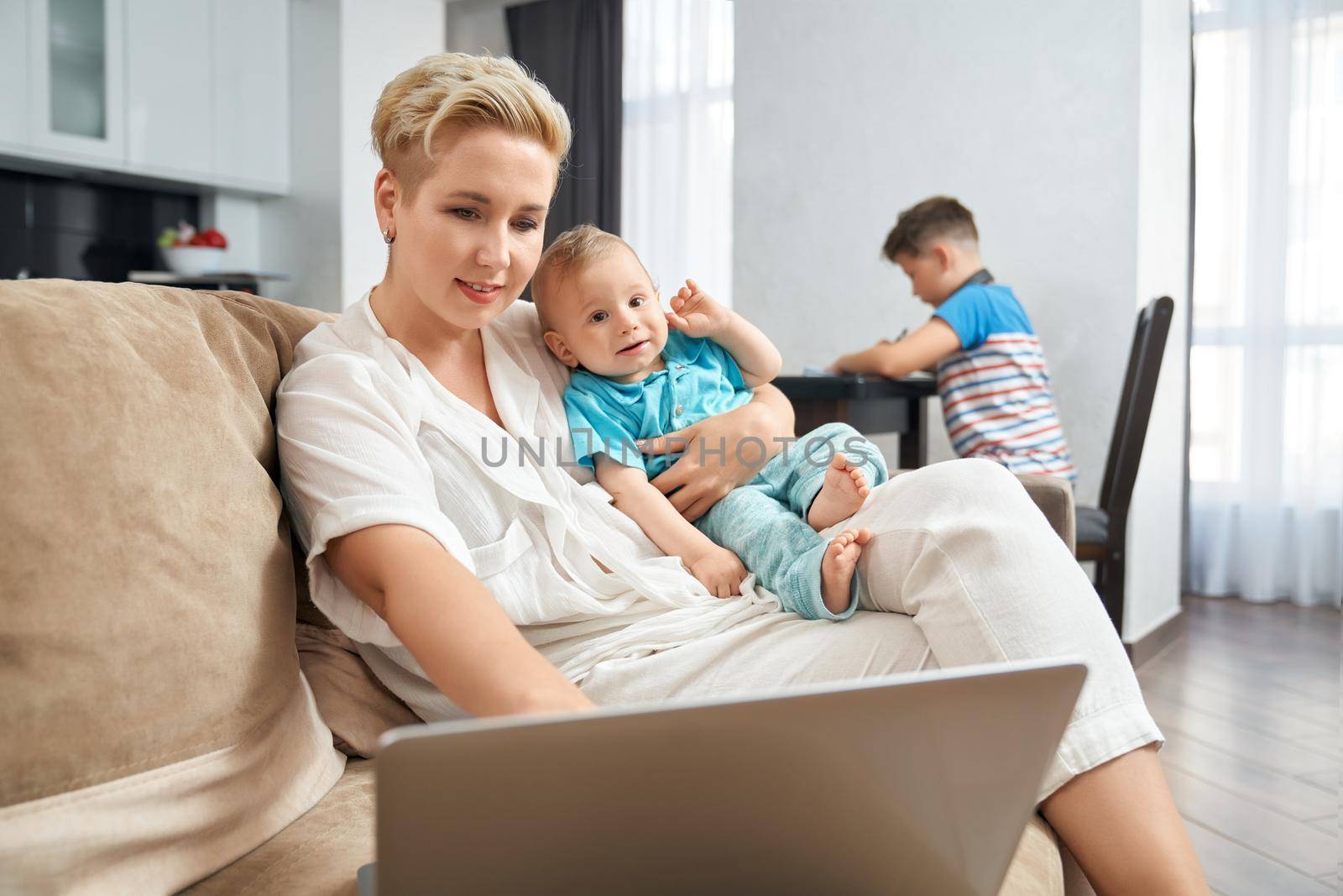 Pleasant woman with blond hair working on laptop and carrying toddler on hands. Blur background of teenage boy sitting on table and doing homework.