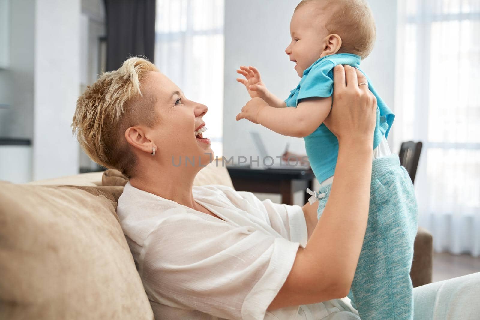 Side view of cheerful woman sitting on cozy sofa and playing with her cute little son. Smiling mother enjoying time spending with her toddler.
