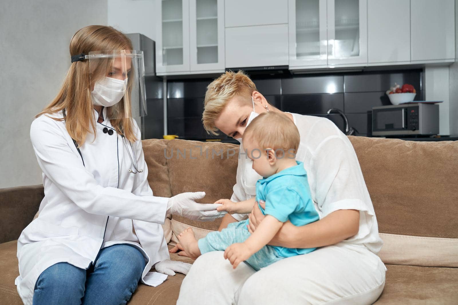Competent pediatrician in protective mask and gloves providing health treatment for toddler that sitting on mother knees. Concept of children medical care.
