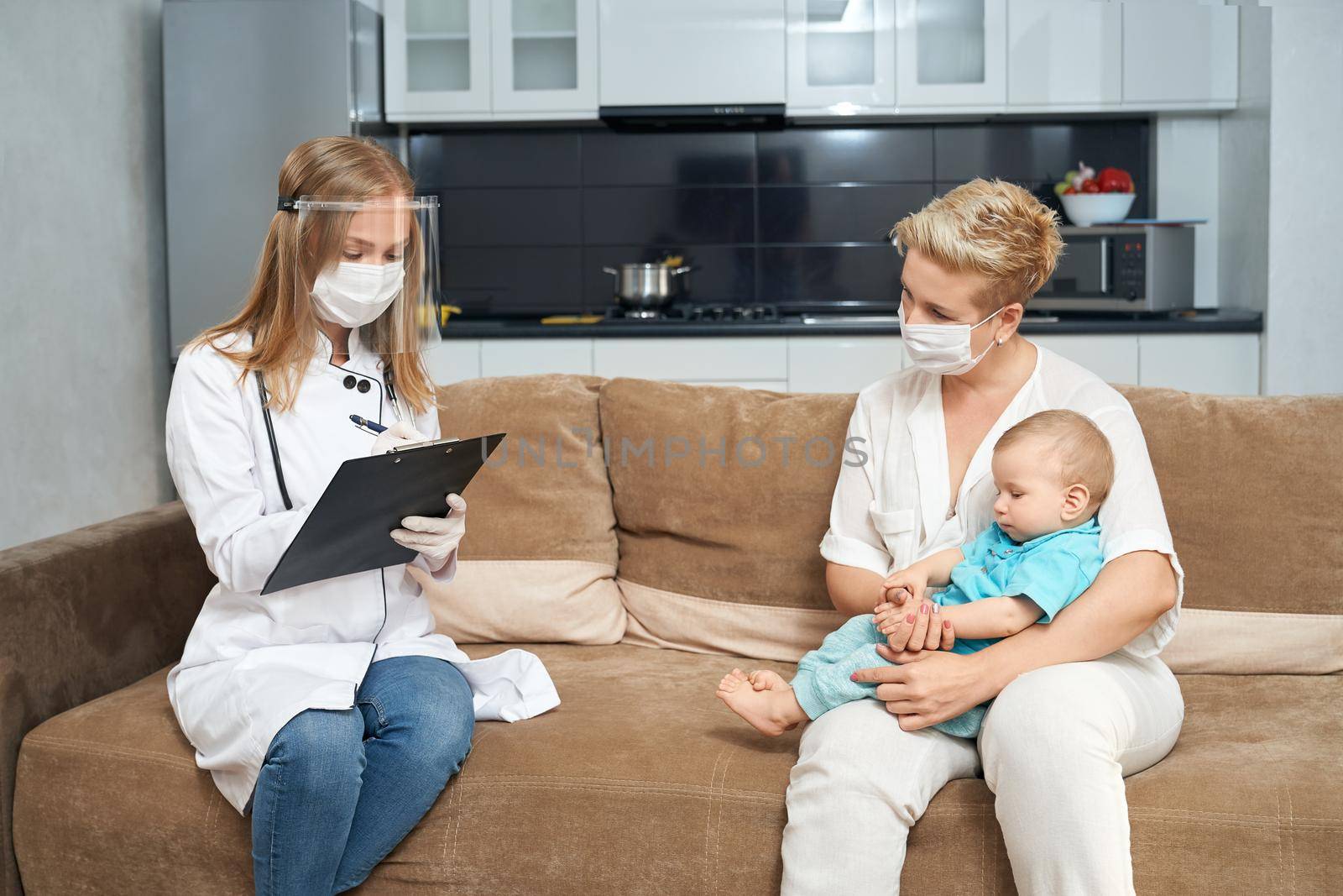 Pleasant woman in medical mask sitting on couch with her little son on knees while female doctor writing on clipboard near. Health care for children at home.