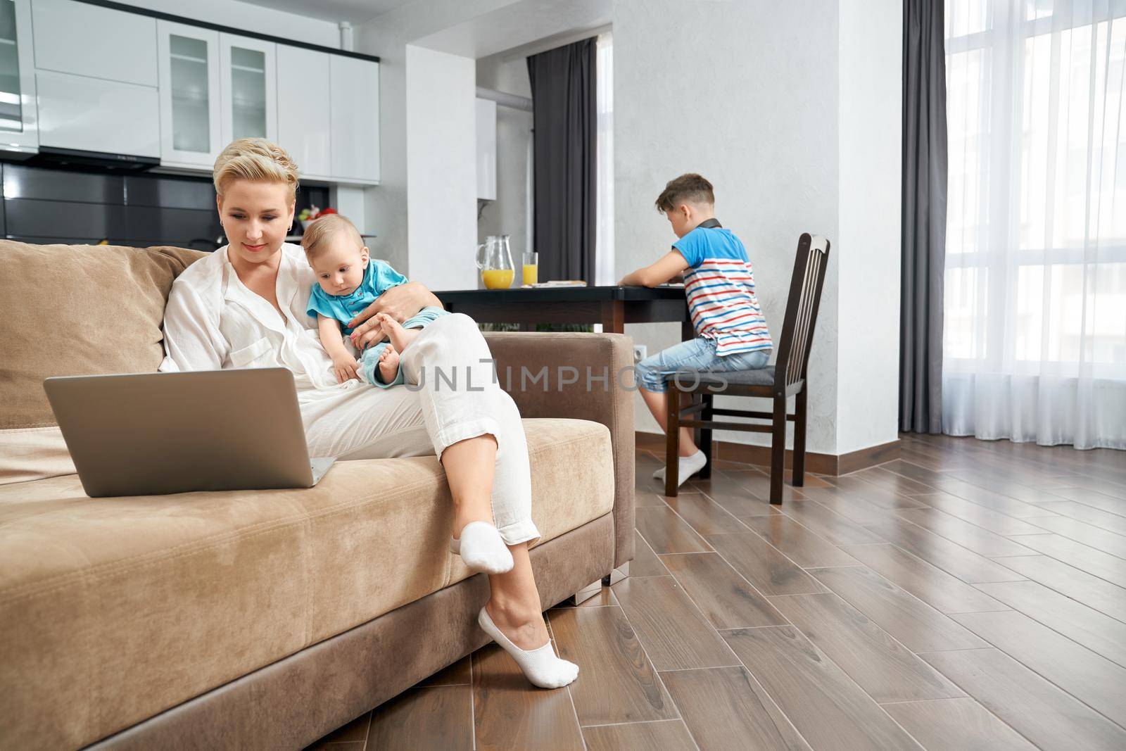 Mother holding baby while sitting on couch with laptop by SerhiiBobyk