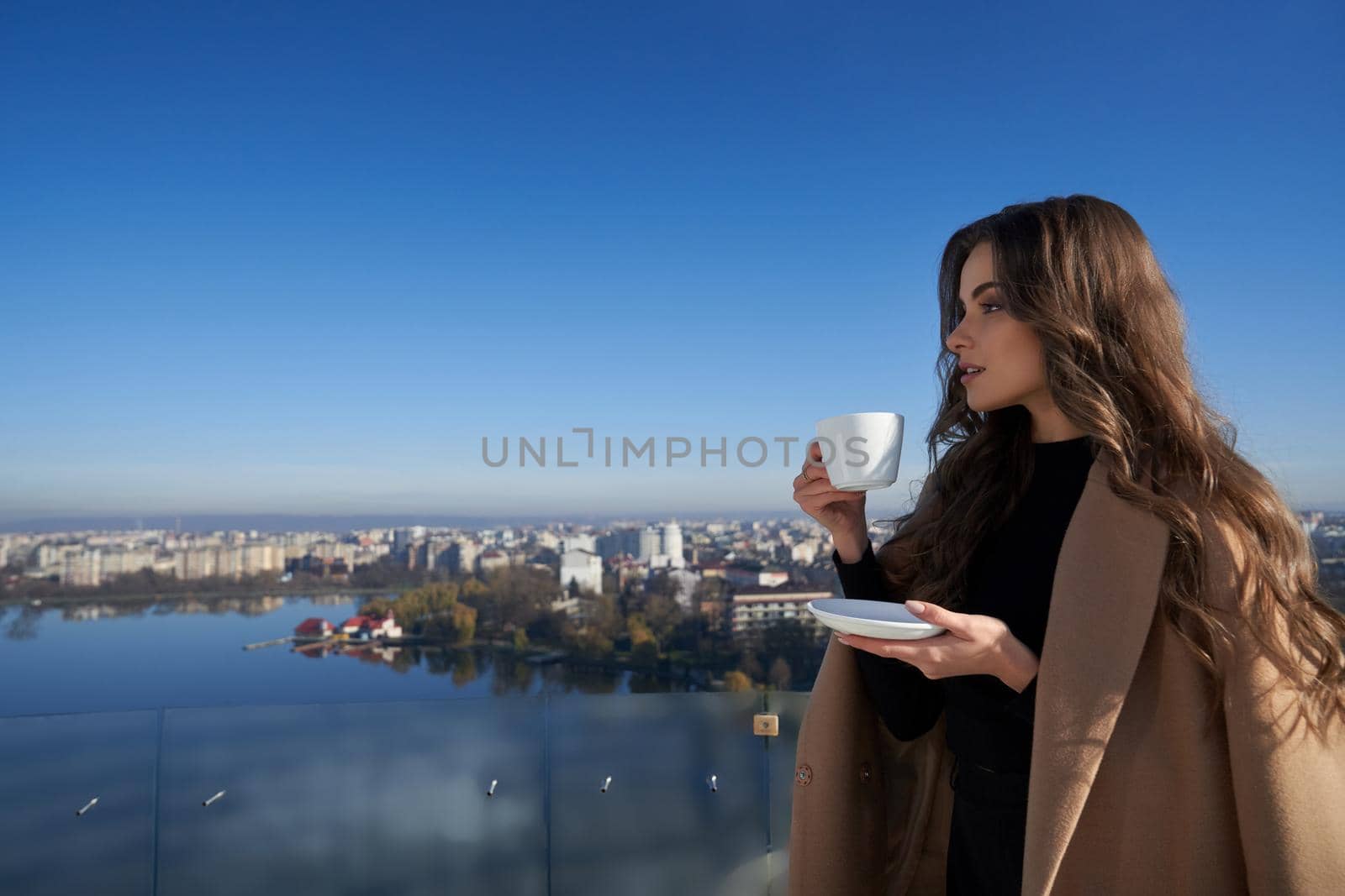 Close up portrait of beautiful young brunette woman standing on balcony with cup coffee or tea and looking on city. Concept of enjoying beautiful nature and city view.