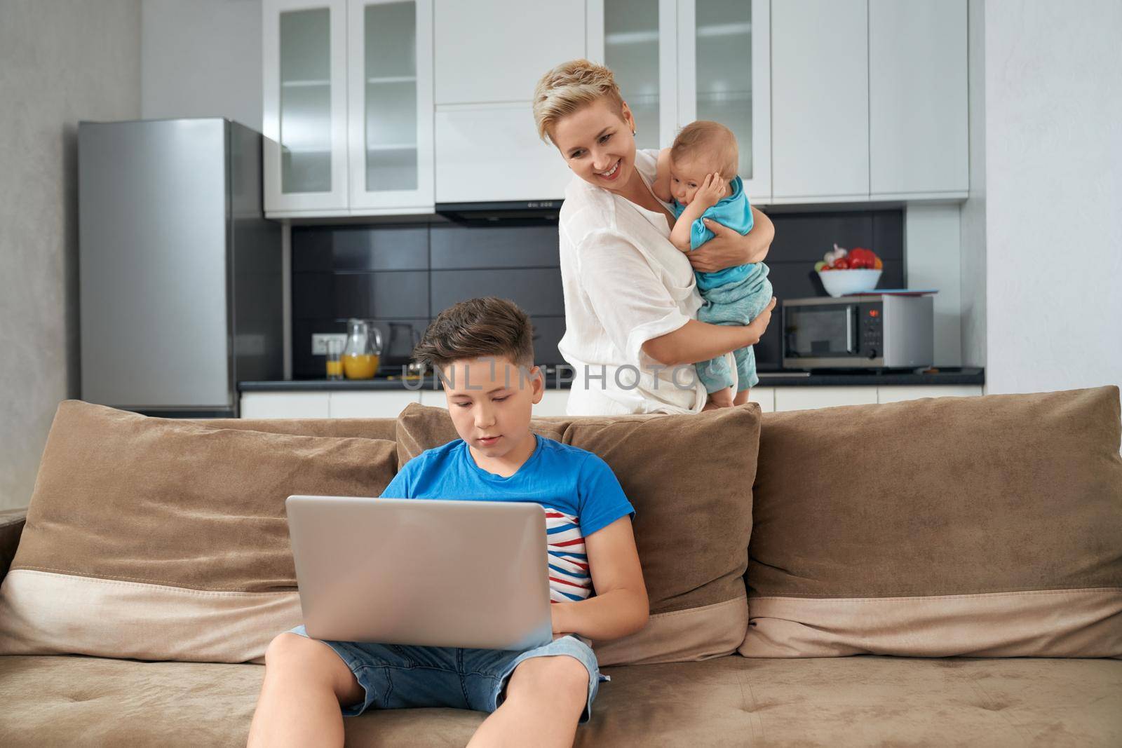 Teenager boy playing games on modern laptop while his mother carrying toddler on hands. Happy family spending time at home. Domestic lifestyle.