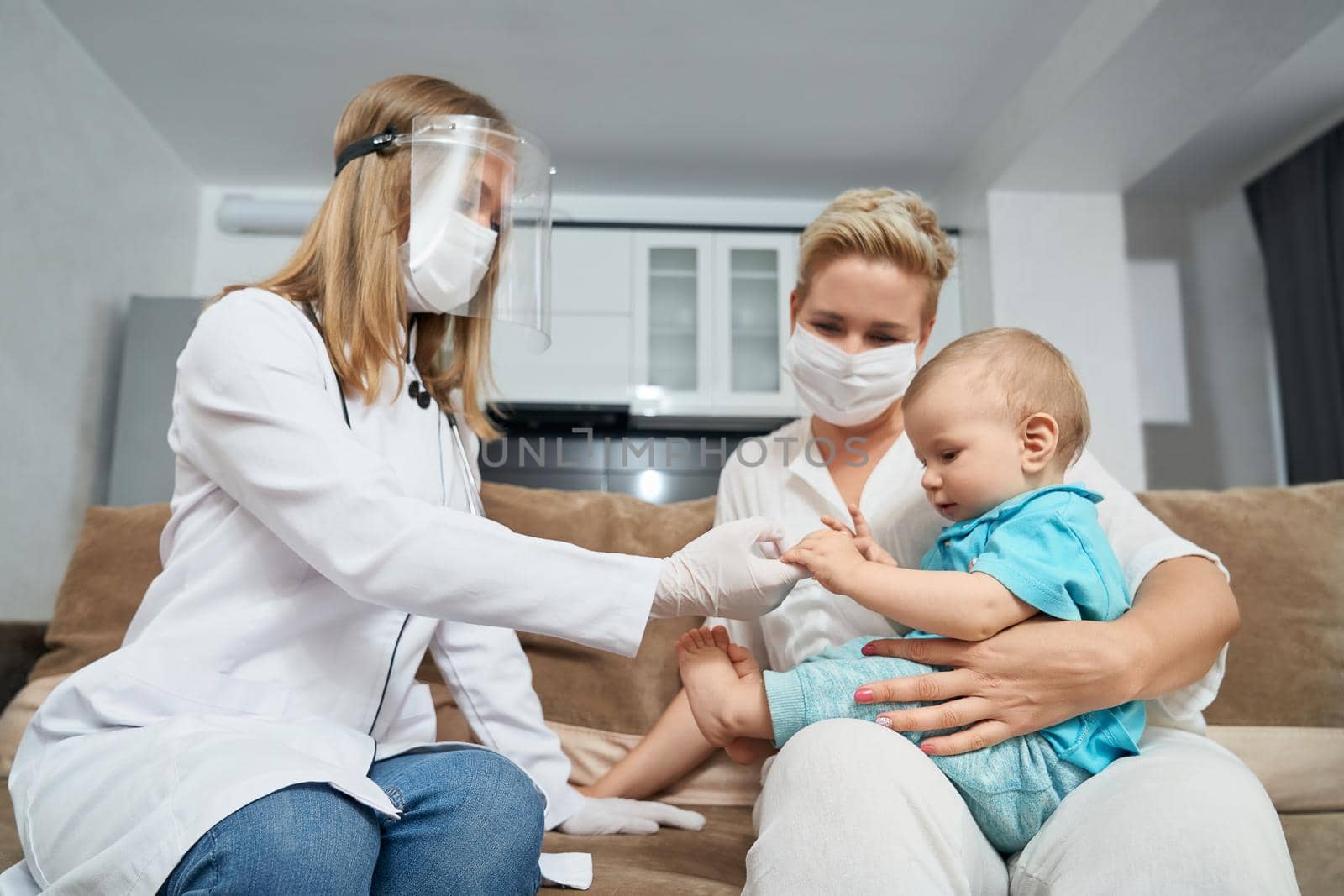 Female doctor in medical uniform and protective mask examining little baby that sitting on mother knees. Nurse checking cute patient at apartment.