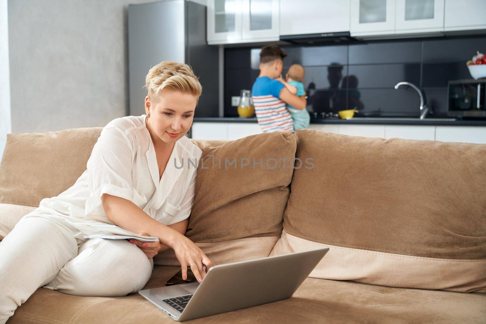 Attractive woman sitting on couch and working on laptop while her eldest son taking care of little baby. Remote work during maternity leave.