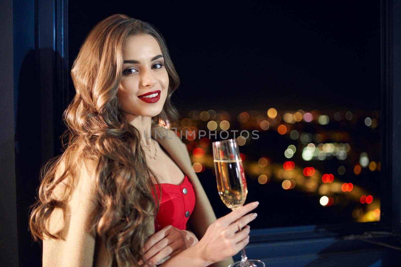 Side view portrait of young beautiful brunette woman standing near window with champagne glasses. Concept of celebration holiday at home with incredible view from window.