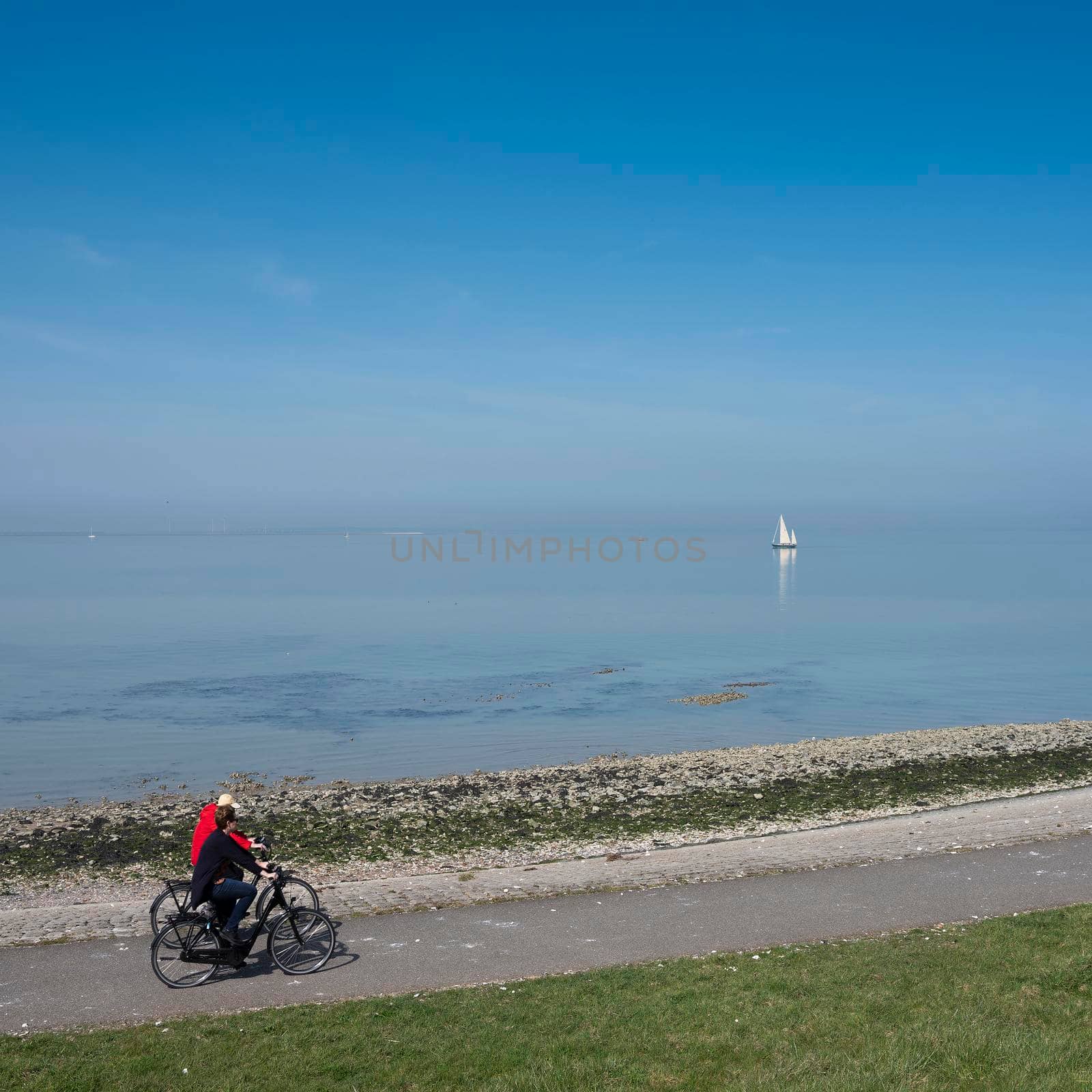 people ride on bicycle track and lonely sailing boat on vast empty blue lake in zeeland by ahavelaar
