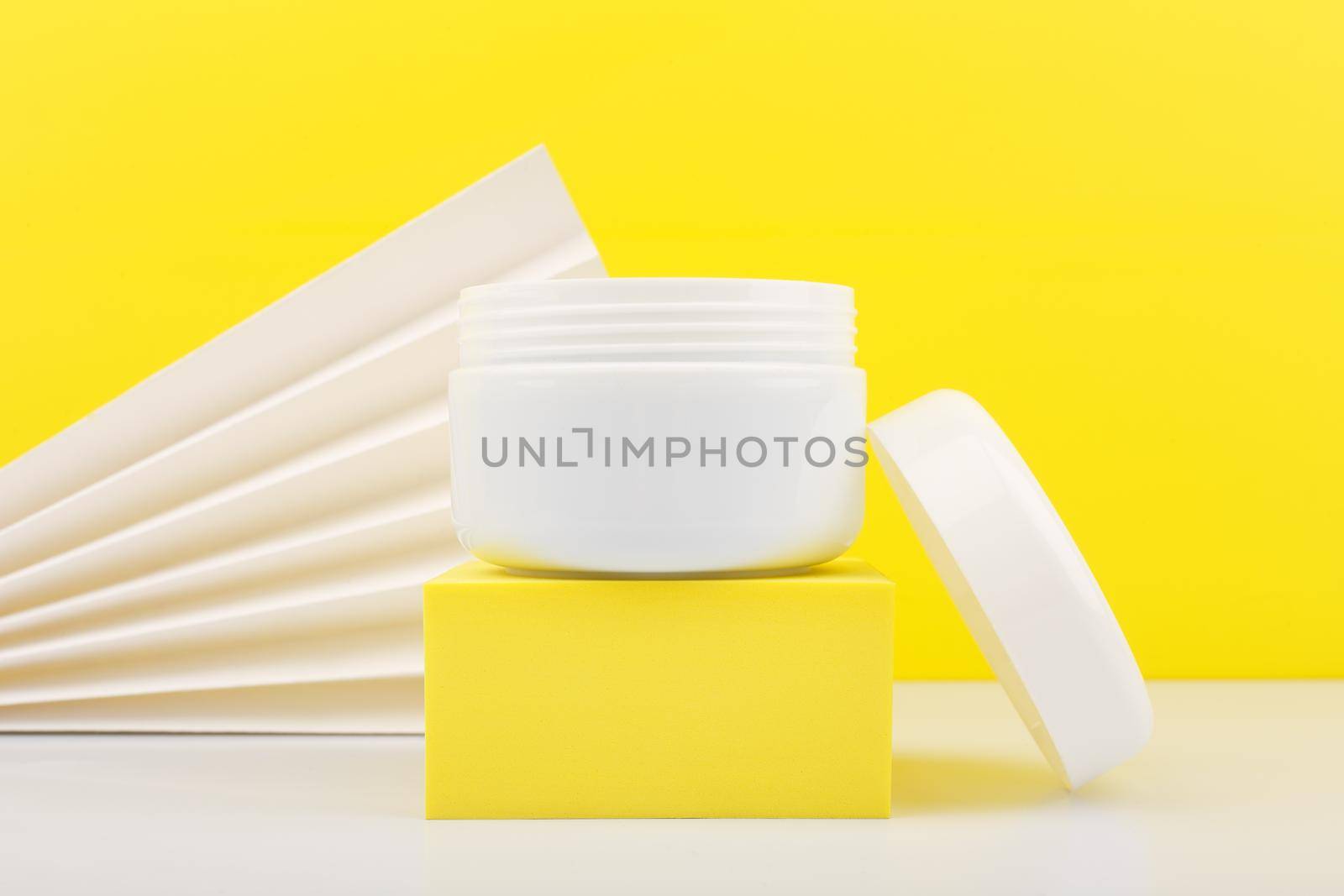 Close up of white opened jar on white table against yellow background decorated with white waver. Concept of summer skin or hair hair with sunblock. Face cream, mask, balm or scrub 