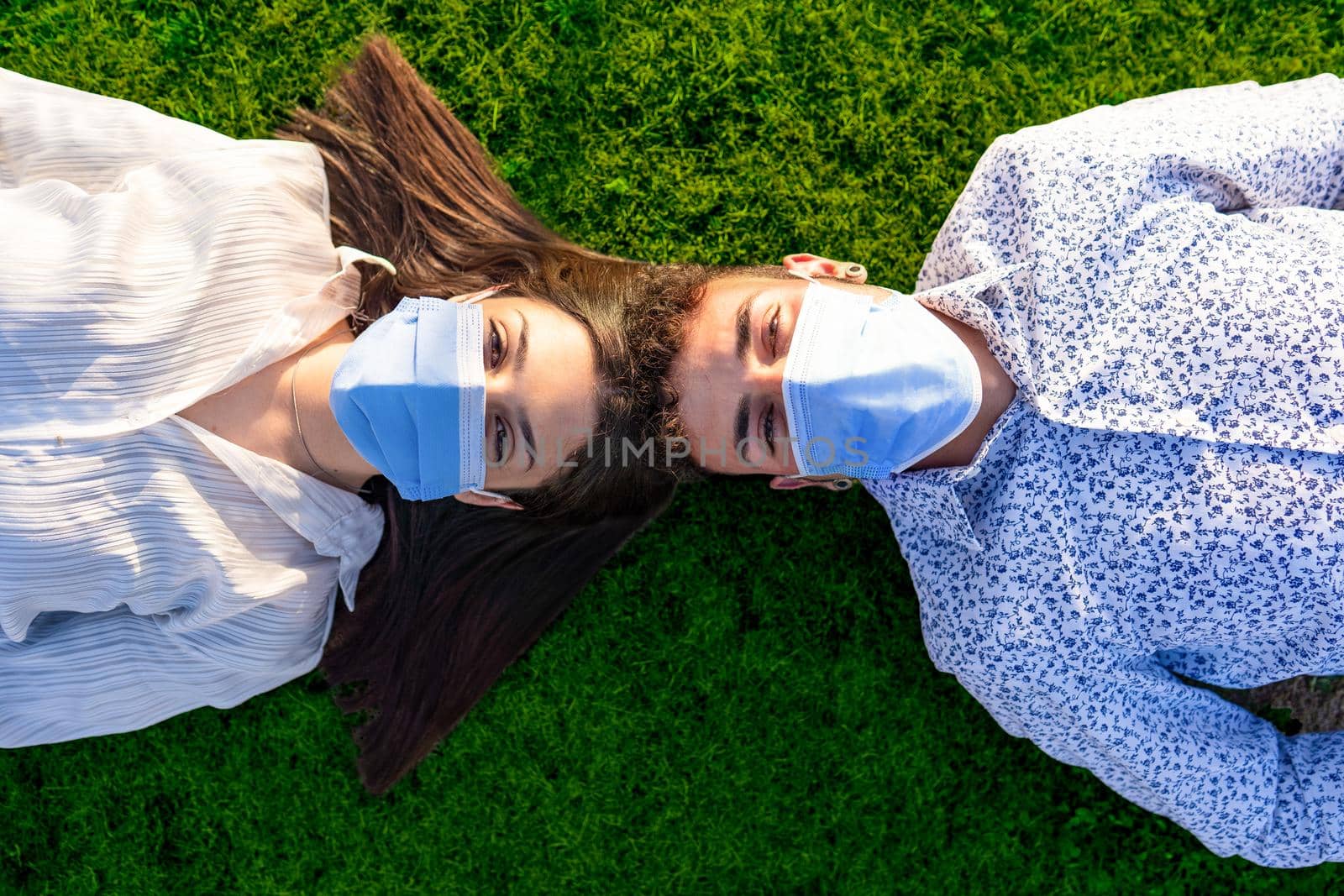 Beautiful couple of young students lying head to head on backs on green lawn of city park looking into the camera wearing the protective mask against the Coronavirus pandemic. New normal health habits by robbyfontanesi