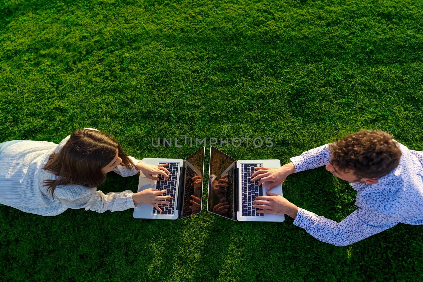 Beautiful couple of young students lying facing each other on lawn grass in city park working on laptop using mobile internet connection. New normal everywhere freelance self entrepreneur job activity by robbyfontanesi