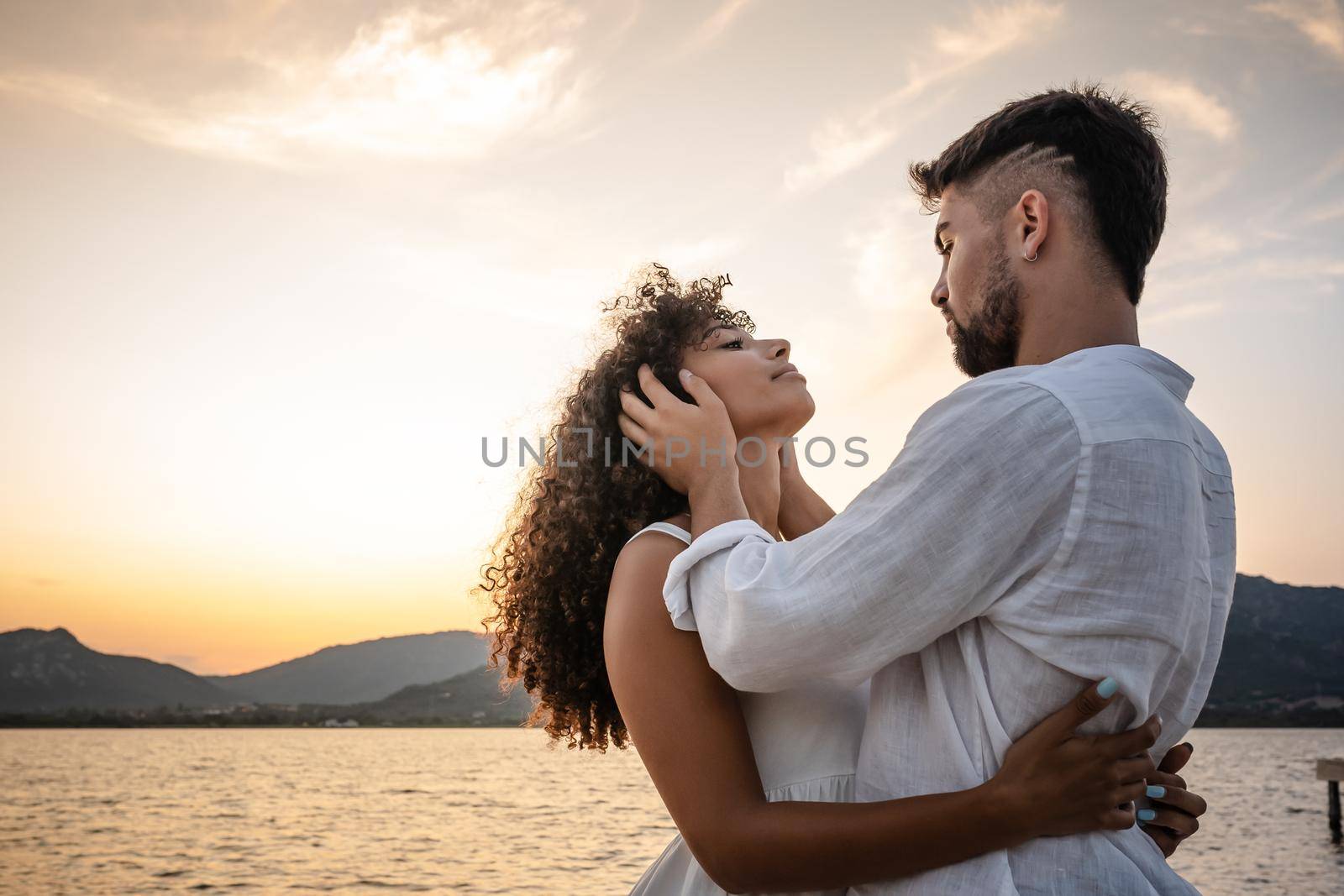 Romantic scene of bearded macho man holding head of his beautiful curly African-American woman in his hands looking into her eyes. Sentimental scene of young couple in love at sunset in nature by robbyfontanesi