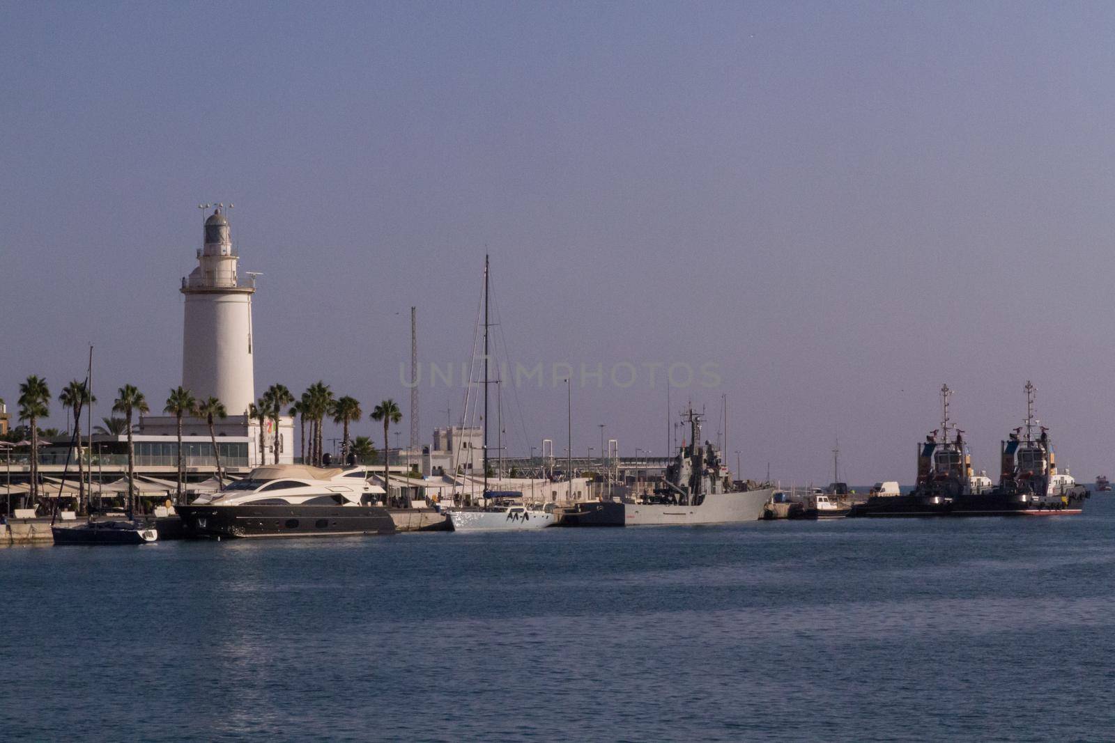 Wide-shot of the dock in a port with siailing boats moored and a white lighthouse