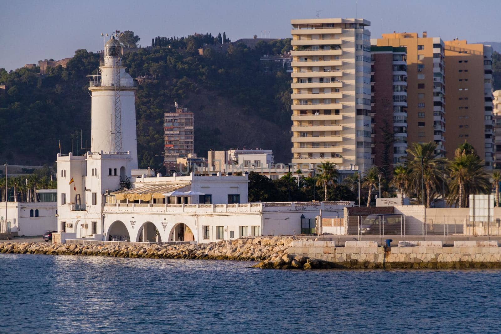 Wide-shot of Malaga's Port Lighthouse on a sunny day with Gibralfaro Castle in the background