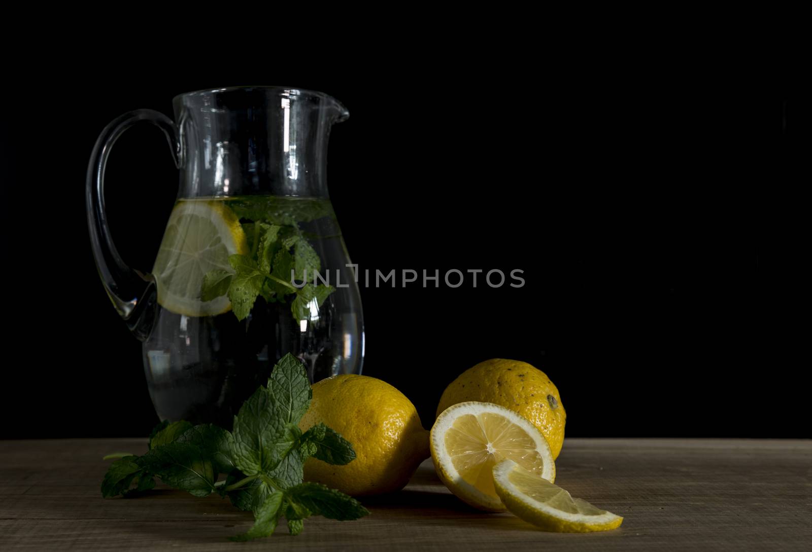 still life with a jug of water filled with mint and lemon with the raw lemon fruit in the foreground on wooden table