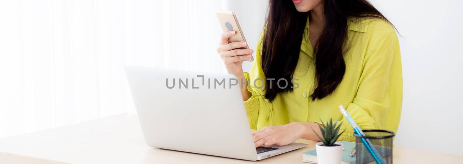 Young asian businesswoman working on laptop computer online on desk at home office, freelance woman looking message on smart phone and typing on notebook on table, business and communication concept. by nnudoo